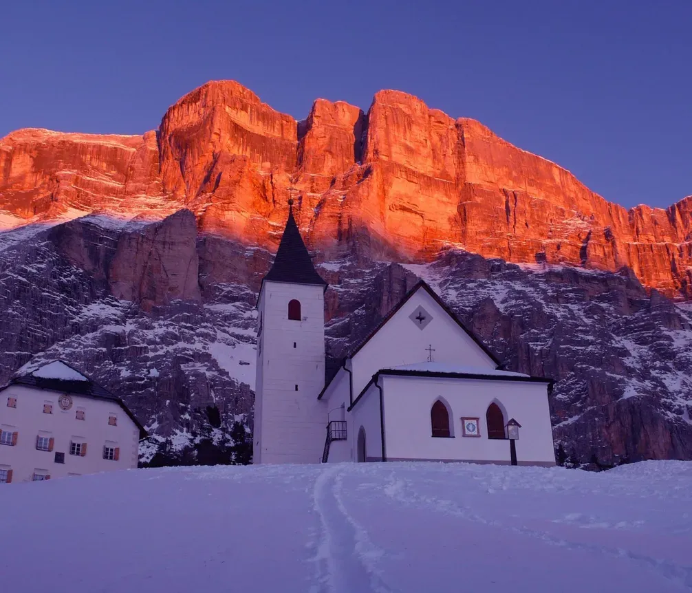 Eine weiße Kirche mit dunklem Dach und Glockenturm vor einer rot-orange leuchtenden Felswand im Dolomiten-Gebirge bei Sonnenuntergang, umgeben von verschneitem Gelände.