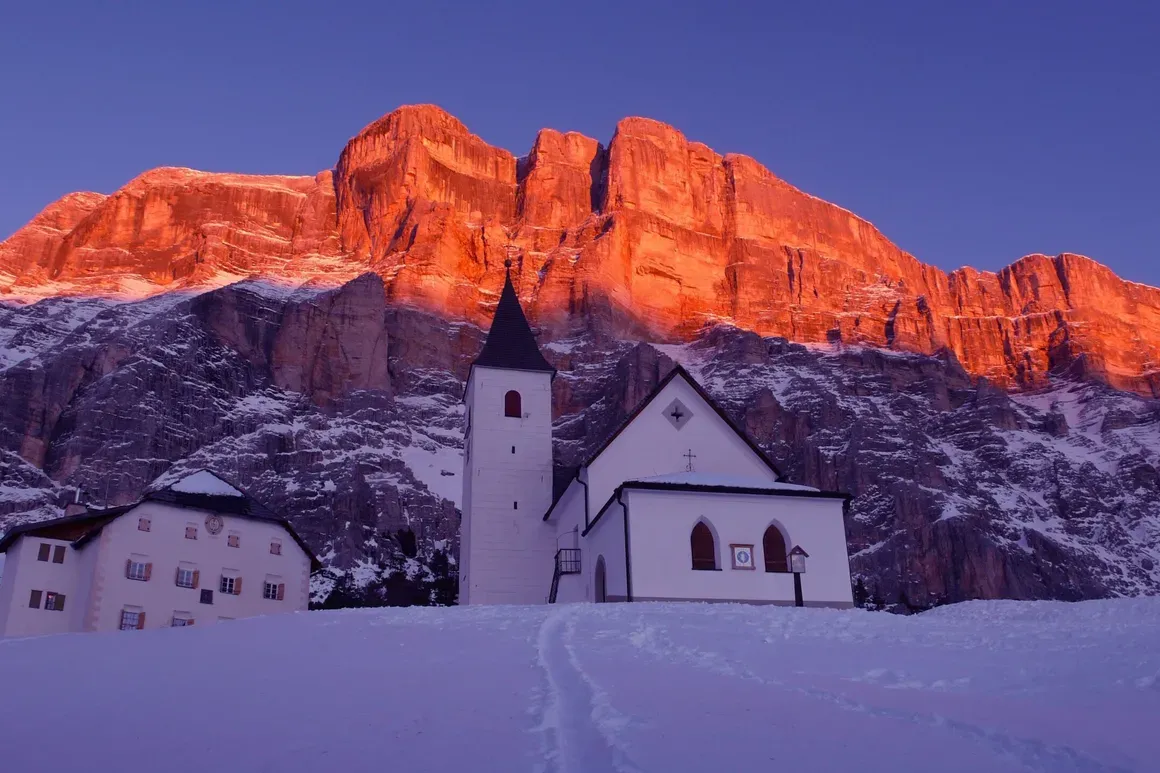 Chiesetta in montagna con pareti rocciose al tramonto, neve sul terreno e cielo azzurro.