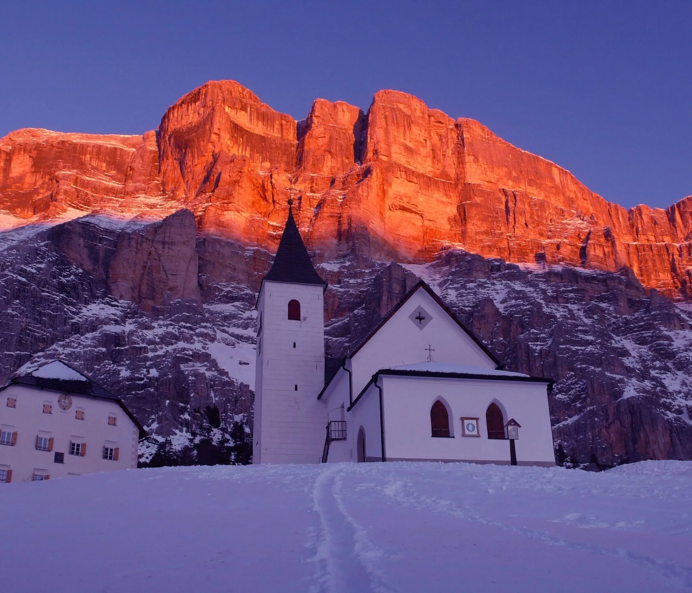 Eine weiße Kirche mit dunklem Dach und Glockenturm vor einer rot-orange leuchtenden Felswand im Dolomiten-Gebirge bei Sonnenuntergang, umgeben von verschneitem Gelände.