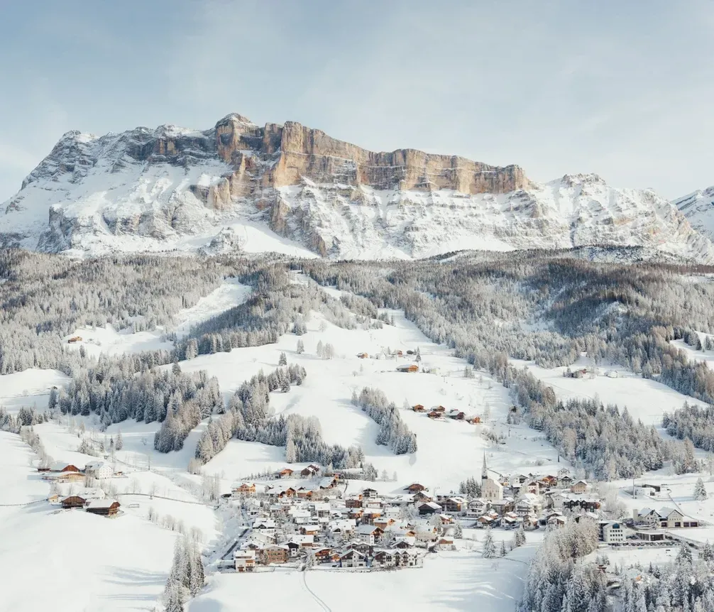 Schneebedeckte Berge in den Dolomiten mit einem kleinen Dorf am Fuße, umgeben von verschneiten Wäldern und Hängen.
