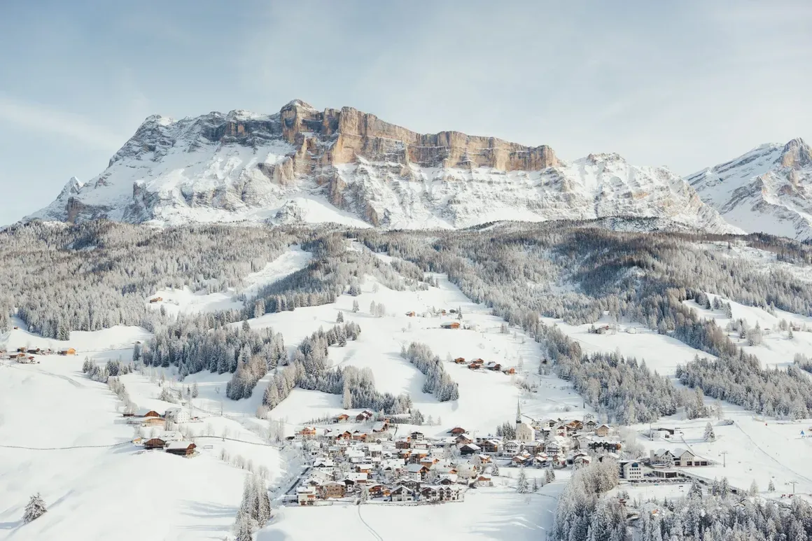 Paesaggio invernale con un piccolo villaggio innevato ai piedi di montagne rocciose e alberi ricoperti di neve, sotto un cielo chiaro.
