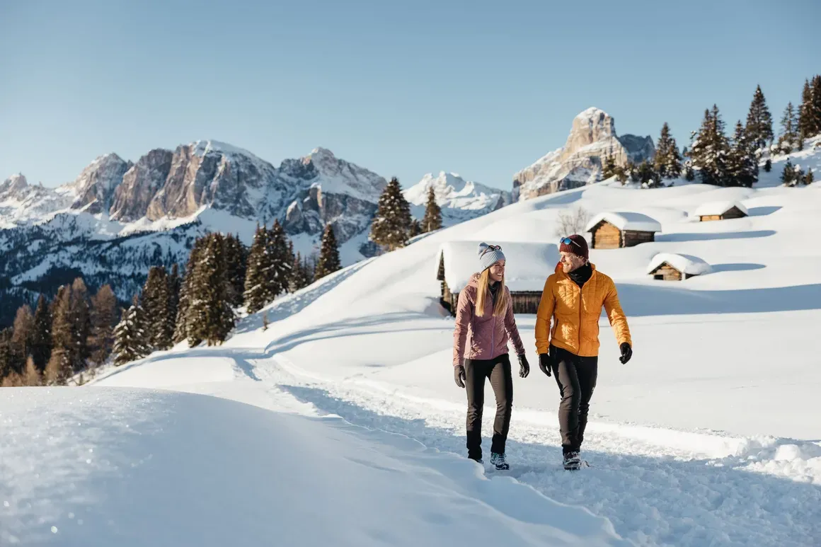 Due persone che camminano sulla neve con montagne e baite sullo sfondo, in una giornata soleggiata nelle Dolomiti, Alta Badia.