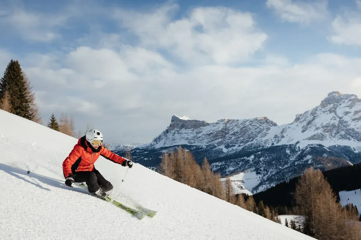 Sciatore in discesa sulla neve con montagne e alberi sullo sfondo, cielo parzialmente nuvoloso, in Dolomiti alta Badia.