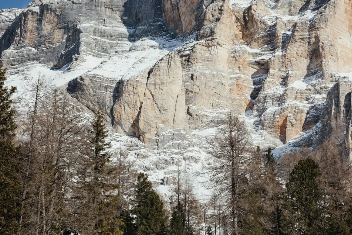 Due sciatori in corsa sulla neve, con alte scogliere rocciose e alberi spogli sullo sfondo, sotto un cielo blu limpido nelle Dolomiti.
