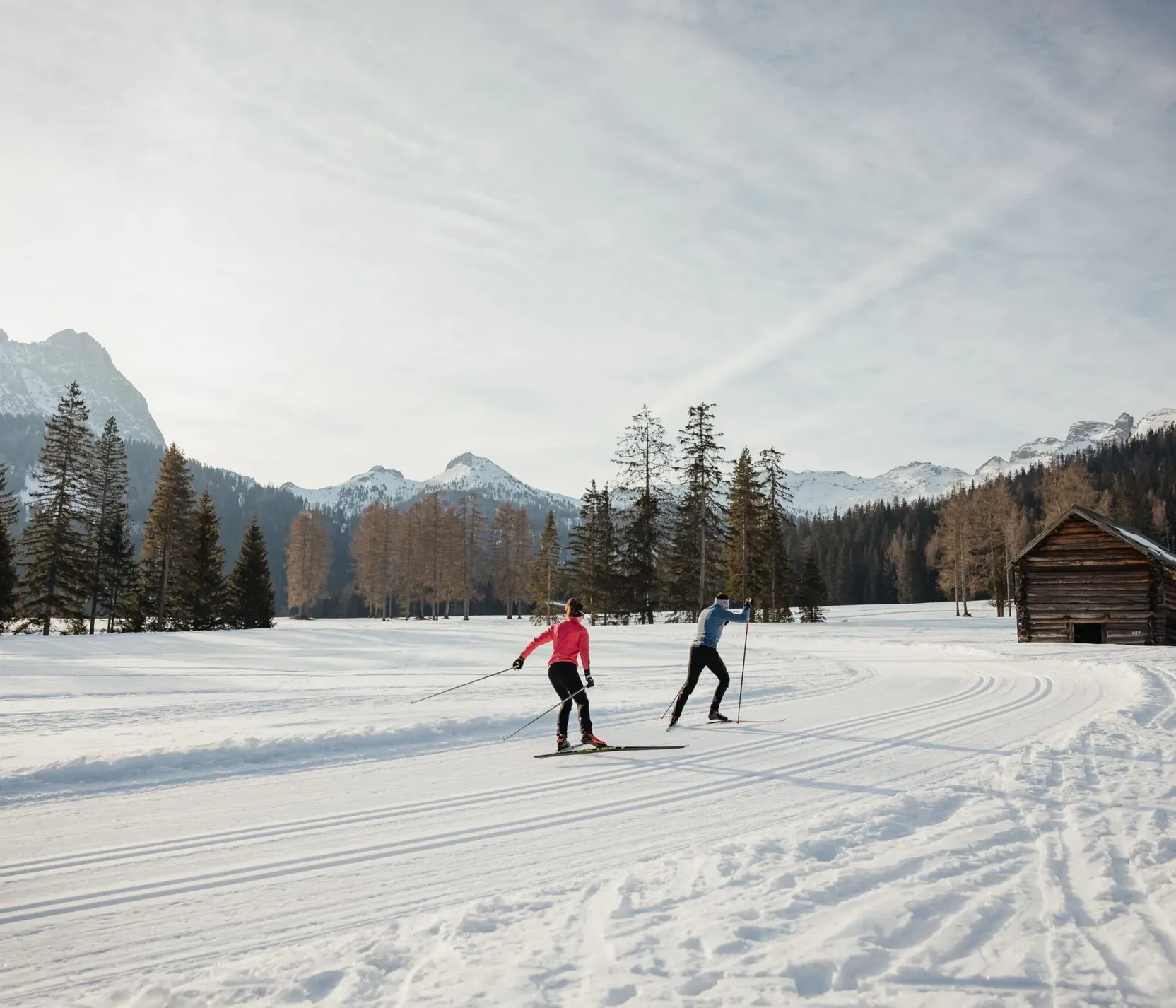 Zwei Skifahrer auf Schneefeld vor Baumgruppe und Bergen in Alta Badia, Dolomiten, sonniger Tag, im Hintergrund eine kleine Holzhütte.