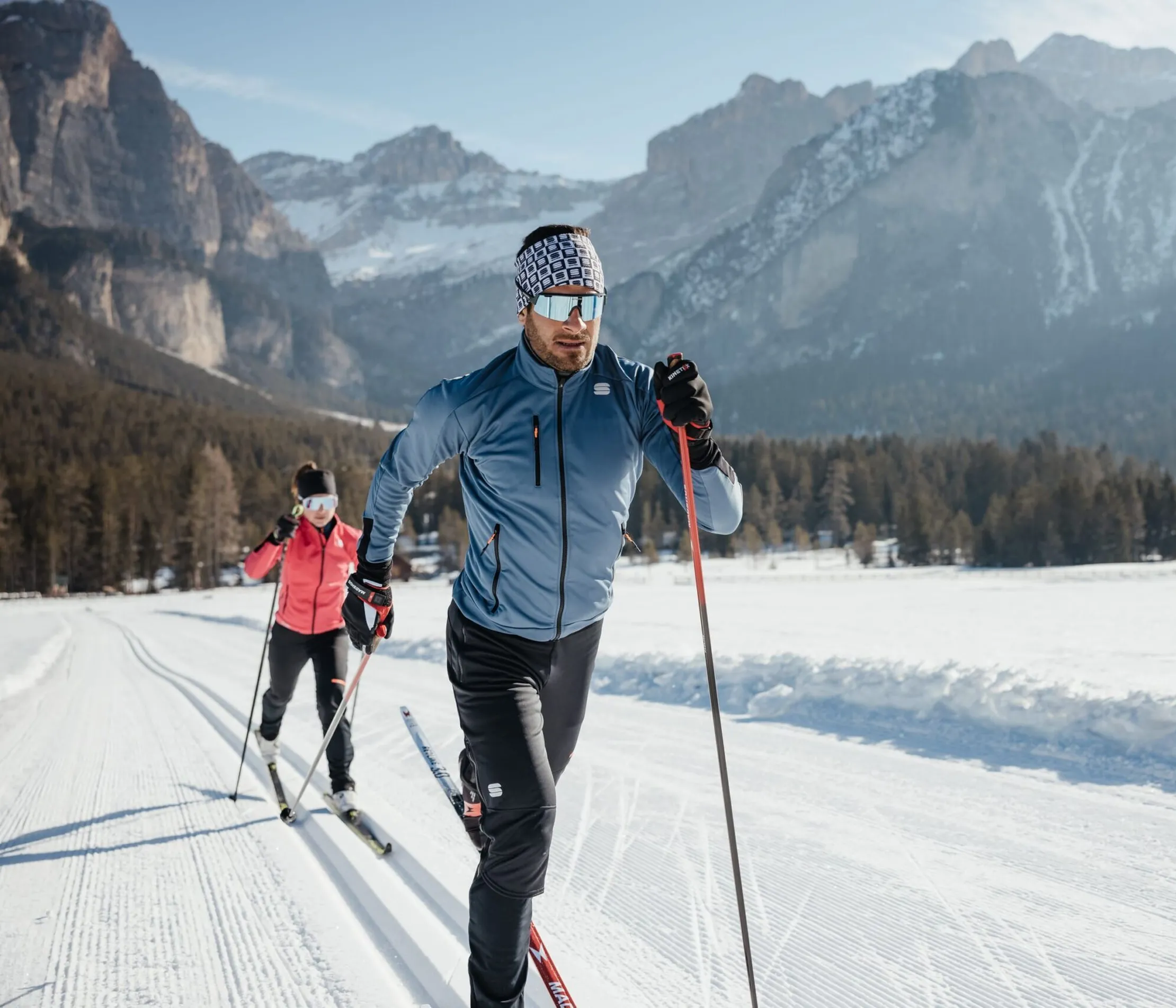 Zwei Skitourengänger in Winterkleidung laufen auf einer präparierten Schneespur in den Dolomiten. Im Hintergrund sind verschneite Berge und Tannen zu sehen.