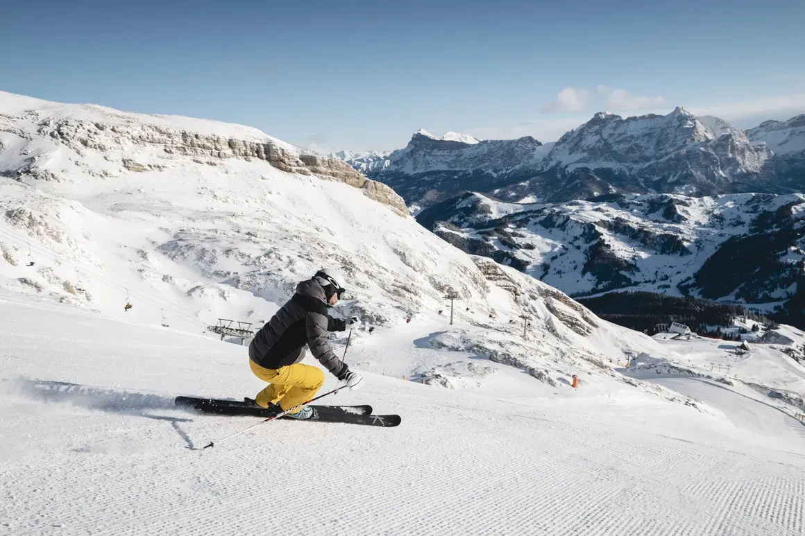 Sciatore in piena discesa sulla neve, con montagne innevate e cielo sereno sullo sfondo nelle Dolomiti di Alta Badia.