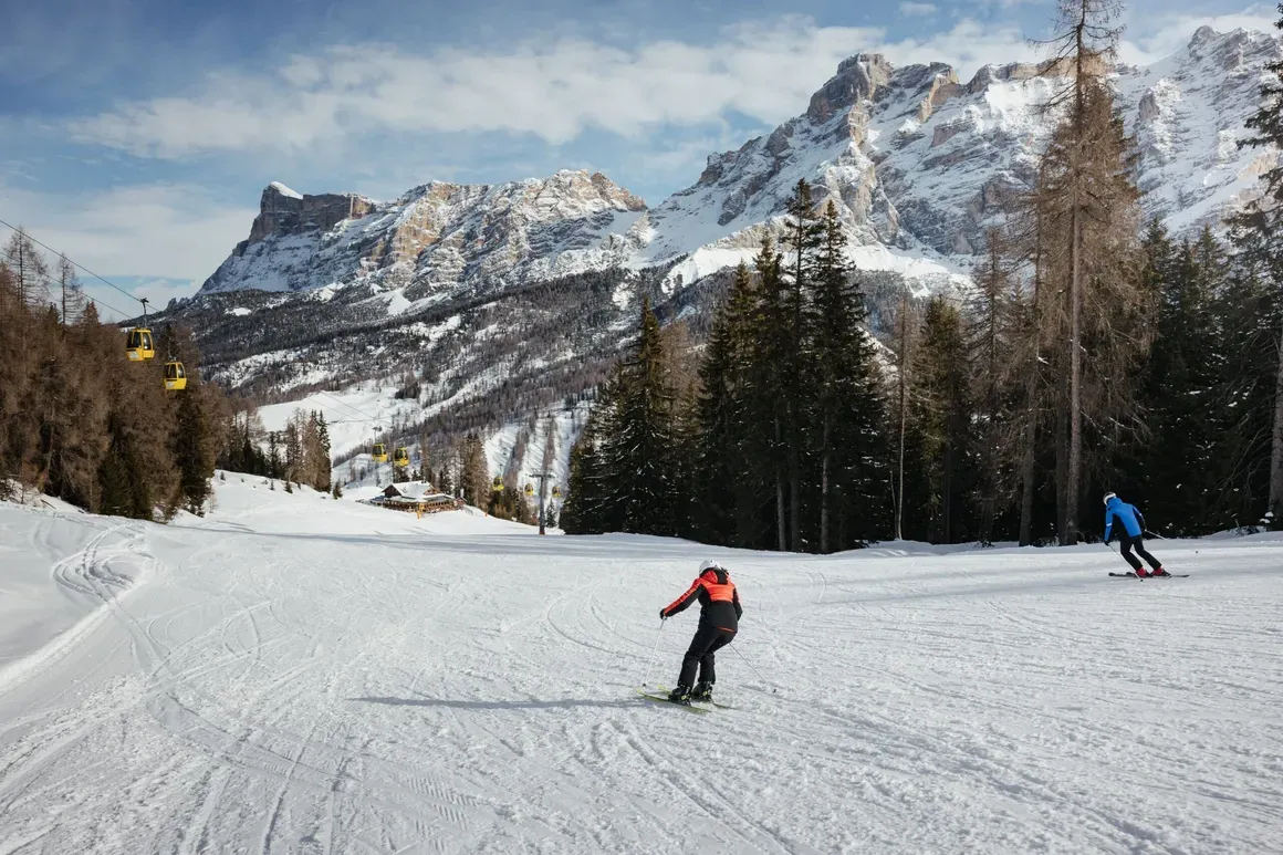 Sciatori che scendono su una pendenza innevata circondata da alberi e montagne con cielo nuvoloso.