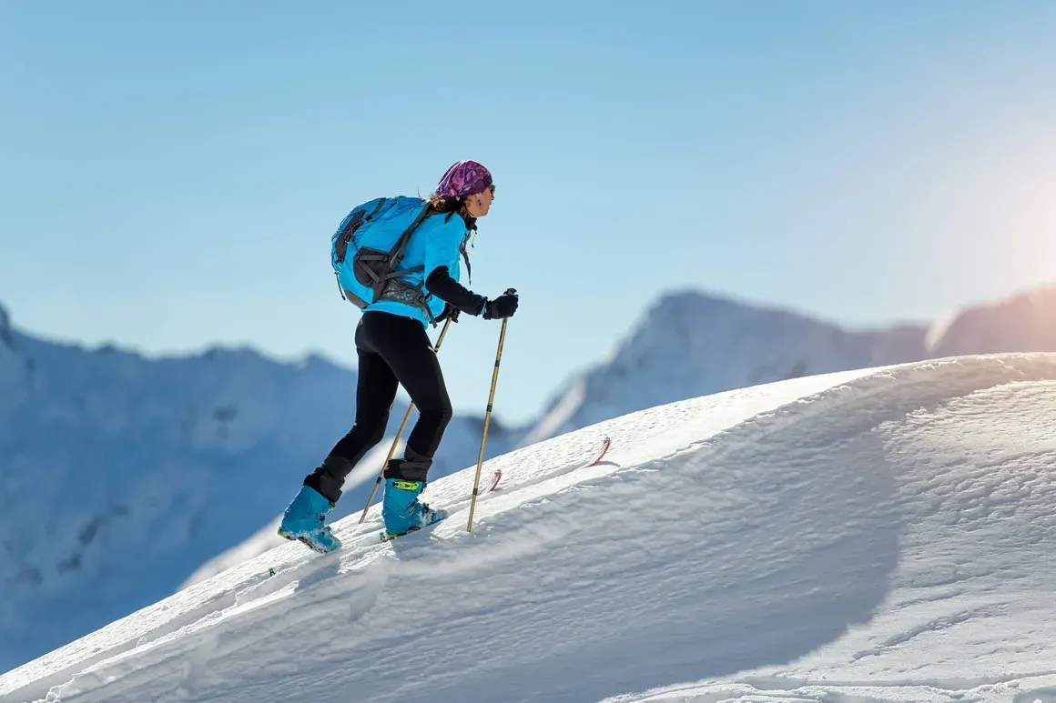 Escursionista in montagna con attrezzatura da sci, sfondo di cime innevate e cielo azzurro, luce solare sulla neve.